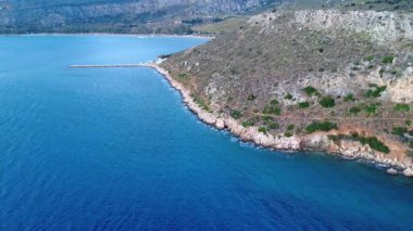 Bir Yunan adasındaki sarp kayalık bir sahil şeridi, dar bir yürüyüş yolunu ve sessiz bir körfezi koruyan bir dalgakıranı gözler önüne seriyor. Akdeniz kıyılarının saf güzelliğini yansıtıyor..