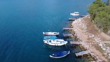 Bu huzurlu Yunan limanında kayalık bir kıyı şeridi boyunca demirlenmiş ahşap balıkçı tekneleri yer alıyor. Tamarisk ve diğer ağaçlar sakin Akdeniz sularında gölge oluşturur..