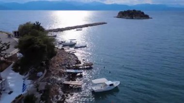 Gün batımında, uzak bir Yunan adasında ahşap kayıklar ve taş rıhtımlarla sessiz bir balıkçı körfezi gösterilir. Limanın yukarısında dalgakıranla korunan ve kayalık bir adanın koruduğu küçük bir köy var..