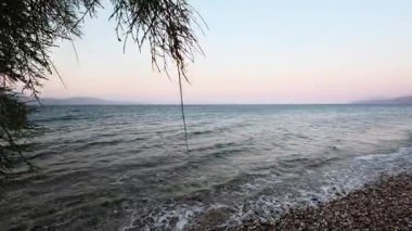 Yunanistan 'da bir çakıl taşı plajı gün batımından önce çekiliyor. Dalgalar ve ağaçlar suyu kaplıyor. Akdeniz manzarası sessiz ve manzaralı..