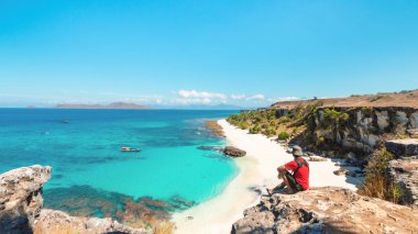 Genç bir adam güneşli bir günde beyaz kumsal ve mavi denizin tadını çıkarırken kayalıklarda oturuyor. Kangge Adası, Alor, Doğu Nusa Tenggara - Endonezya