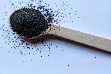 Ahşap bir kaşıkta saf beyaz arka planda kokulu siyah haşhaş tohumları. Doğal ışıklandırma ile minimalist kompozisyon tohum dokusu ve zengin rengi vurgular.