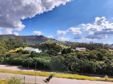 Yeşil dağları ve beyaz bulutlarla benekli mavi gökyüzünü kaplayan güzel bir panoramik manzara. Manzara, arka planda ağaçlar ve ihtiyatlı bir bina ile çevredeki yemyeşil bitki örtüsünü gözler önüne seriyor. Bu doğal ortam huzuru çağrıştırıyor.