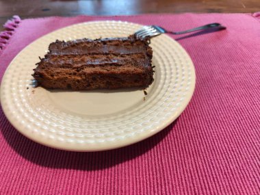 Lezzetli bir dilim çikolatalı pasta kremalı bir tabakta servis ediliyor, yanında bir çatal, canlı pembe bir masa örtüsü, tatlı ve samimi bir anı çağrıştırıyor..