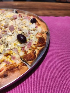 Peynir, soğan, zeytin ve mısırın karışımı ile kaplanmış, ağzınızı sulandıran altın hamurlu gurme pizzanın yakın çekim görüntüsü. Pizzanın bir dilimi canlı pembe bir masa örtüsünün üzerinde, kabuğun dokusunu ve taze malzemelerini gözler önüne seriyor.