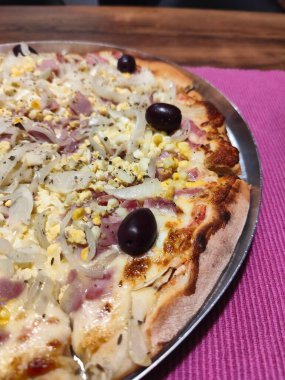 Jambon, soğan, siyah zeytin ve üzerine mısır serpilmiş taze pişmiş bir pizzanın ağız sulandırıcı yakın çekimi, lezzetli bir tat ve doku karışımı sergiliyor..