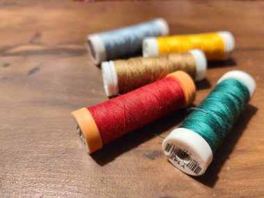 Kırsal ahşap bir masada ustalıkla düzenlenmiş altı renkli dikiş ipliği makarasından oluşan canlı bir koleksiyon. Makarnalarda kırmızı, yeşil, altın ve mavi de dahil olmak üzere çeşitli renkler yer alıyor, kumaşların güzelliğini ve dikiş işçiliğini gözler önüne seriyor. 