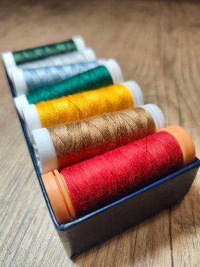 Kırsal ahşap bir zemine karşı siyah bir kutuda düzgünce düzenlenmiş canlı bir iplik makarası koleksiyonu. Spekülasyonlar yeşil, gümüş, sarı, altın, kahverengi ve kırmızı gibi çeşitli renklerde gelir.