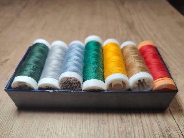 Ahşap bir masa arkasına yerleştirilmiş yeşil, gümüş, altın ve kırmızı tonlarda dahil olmak üzere çeşitli renklerde, canlı dikiş ipliklerinden oluşan organize bir koleksiyon. İplikler, dünya ile güzel tezat oluşturan zarif bir mavi kutuda saklanıyor.