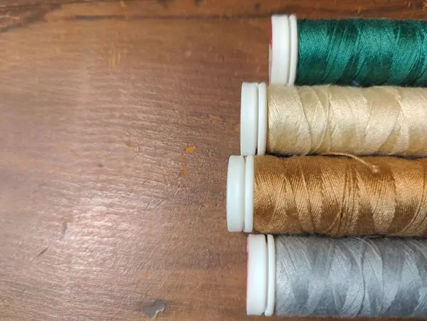 Tahta bir yüzey üzerinde dört adet canlı dikiş ipliği düzgünce düzenlenmiştir. İplikler yeşil, altın, bronz ve gümüşün tonlarında, çeşitli zanaat projeleri ve dikiş işleri için uygun zengin doku ve renkleri gösteriyor.