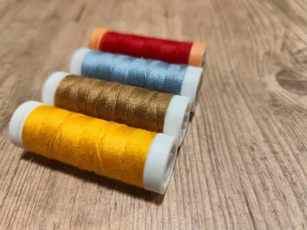 Sarı, altın, mavi, kırmızı ve turuncu renkleri içeren bir renk spektrumunu gösteren canlı dikiş makaralarının organize bir gösterisi. Spikerler düzgün bir şekilde sade ahşap bir yüzeye yerleştirilmiş, ipliklerin dokusunu ve ahşap tahılını vurguluyor,