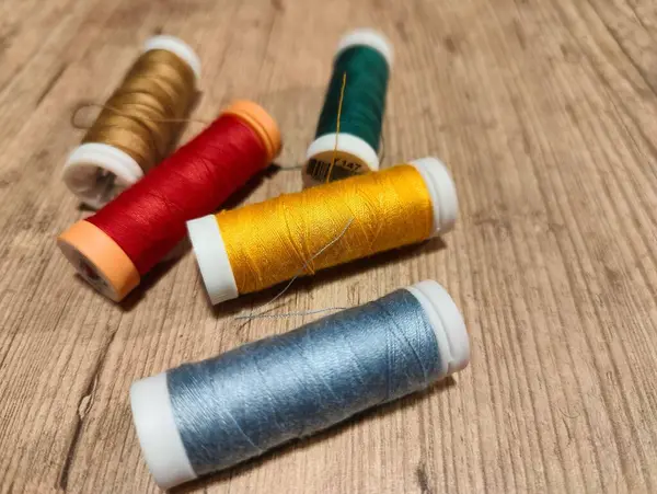 Kırmızı, altın, yeşil, turuncu ve mavi gibi çeşitli renklerde, ahşap bir yüzeye saçılmış, canlı bir dikiş ipliği koleksiyonu. Düzenleme, kumaşların ve el sanatlarının güzelliğini, yaratıcılığı ve ilhamı davet ediyor..