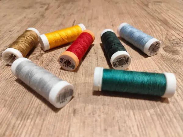 Ahşap bir yüzeye yayılmış çeşitli renkli dikiş ipliklerinden oluşan canlı bir düzenleme. Sahne, altın, sarı, kırmızı, yeşil, açık mavi, turuncu ve gümüş gibi neşeli renklerin bir karışımını yakalar. Çeşitlilik farklı dokuları vurgular