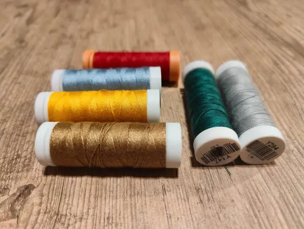 Kırmızı, turuncu, mavi, sarı, altın, yeşil ve gümüşten oluşan çeşitli renklerde, kırsal bir ahşap masaya dikkatlice yerleştirilmiş, canlı dikiş iplikçiklerinin sanatsal bir düzenlemesi. Görüntü her bir çizginin dokusunu gösteriyor, parlak fini rengini vurguluyor.