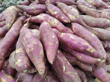 Canlı mor tatlı patates yığını, düzensiz derilerini ve dokularını sergiliyor. Patatesler gelişigüzel dizilmiş, ürünlerin bolluğunu ve tazeliğini gösteriyor, sağlık ve doğallığı çağrıştırıyor..