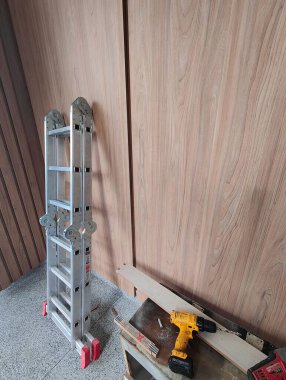 Çalışma alanının bir köşesini, ahşap bir duvara yaslanmış alüminyum bir merdivenle gösteren bir resim, yanında sarı bir matkap ve ahşap kırıntıları, çalışma ve yenileme çalışmaları çağrıştırıyor..