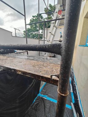 Muşambayla kaplı metal bir yapının yanına yerleştirilmiş bir inşaat merdiveni resmi. Sahne, bulutlu bir günde arka planda ağaçlarla birlikte çalışma ortamında geçen merdivenin paslı metalini vurguluyor..