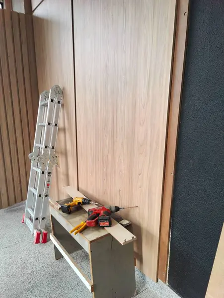 Görüntü, açık renkli ahşap duvarlı bir inşaat alanını gösteriyor. Önde, sürmekte olan bir yenileme projesini vurgulayan tornavida ve merdiven gibi araçlar var.