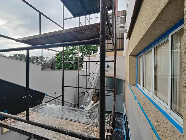 Endüstriyel bir sahnede bina tamiri için kullanılan metal iskeleler görülüyor. Bir işçi aktif olarak resim yapıyor ya da dış görünüşünü koruyor. Bulutlu bir gün, şehir dönüşümünü sembolize eden odaklanmış, yoğun bir çalışma ortamı yaratır..