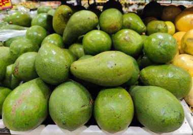 Bir markette sergilenen, mükemmel olgunlukta yeşil avokado yığını. Canlı renkleri ve parlak derileri, sağlık ve vitamin hissini aktararak tazeliği ve kaliteyi vurgular..