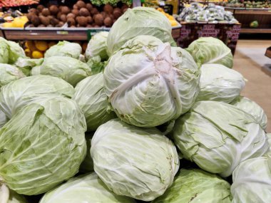 Bir markette yığılmış canlı yeşil lahana gösterisi taze dokularını ve sağlıklı görünüşlerini ön plana çıkarıyor, çeşitli meyve ve sebzelerden oluşan bir zemin üzerine kurulmuş, canlı bir çiftlik havası uyandırıyor..