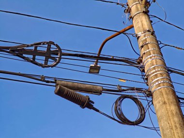Dikey bir sokak lambasının net bir görüntüsü çeşitli elektrik kabloları ve kablolar açık mavi bir gökyüzüne karşı asılıdır. Detaylar keskin, destekleri ve izolatörleri gösteriyor, kentsel elektrik altyapısını vurguluyor.