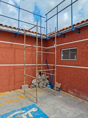 Çalışma alanındaki bir inşaat iskeletinin görüntüsü, dokulu kırmızı bir duvara dayanıyor. Metal iskele merdivenleri ortaya çıkardı ve arka planda bir pencere görülebiliyordu. Sahne, devam eden çalışma ve inşaat faaliyetlerini aktarıyor, materyalleri vurguluyor