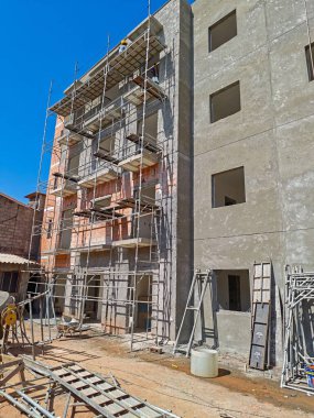 İnşaat halindeki çok manzaralı bir binayı gösteren bir görüntü, etrafında iskeleler, beton duvarlar ve açık mavi bir gökyüzüne karşı korunmasız tuğla yapı. Sahne, inşaattaki ilerleme ve sıkı çalışmayı gösteriyor..