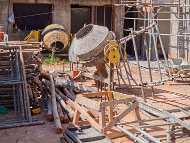 Arka planda ve iskelelerde rastgele yerleştirilmiş metal borularla çevrili beton karıştırıcı gibi ekipmanlarla dolu canlı ve hareketli bir inşaat alanı. Güneş malzemelerin kırsal dokusuna vurgu yapıyor ve bir atmosfer yaratıyor.