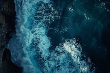 Okyanus dalgalarının karanlık ve kayalık bir kıyıya karşı kırılışlarını gösteren bir yukarıdan çekim. Görüntü, derin deniz suyuyla çarpışan dalgaların parlak beyaz köpüğü arasında çarpıcı bir kontrastı gösteriyor.
