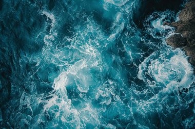 Engebeli bir kıyı şeridine doğru dalgalanan okyanus dalgalarının güçlü enerjisini yukarıdan yansıtan bir perspektif. Canlı deniz mavisi suları beyaz başlıklar ve köpüklerle noktalanarak dinamik ve dokusal bir görüntü oluşturur. T 'nin koyu mavisi arasındaki kontrastlık