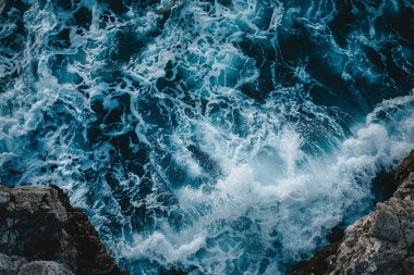 Engebeli bir kıyı şeridine doğru dalgalanan okyanus dalgalarının dinamik enerjisini nefes kesen bir bakış açısı yakalar. Suyun koyu mavi renkleri, çarpan dalgaların beyaz köpüğüyle çok güzel tezat oluşturuyor ve görsel olarak çarpıcı bir görüntü oluşturuyor.
