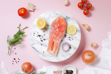 Yukarıdan bakıldığında, limon dilimleri, sarımsak, otlar, baharatlar, domatesler, soğanlar ve beyaz kumaş aksanlarıyla mermer bir tabakta somon bifteğini pastel pembe arka planda gösteriyor..
