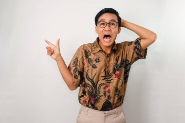 Üzgün yüzlü, batik ve gözlüklü, el hareketleriyle yan tarafı işaret eden Asyalı bir adam.