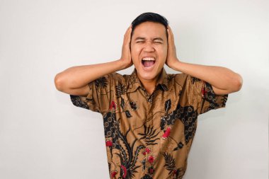 İfade bozukluğu olan Asyalı bir adam, batik giyiyor, iki eli de kulaklarını kapatıyor.