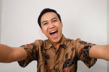 Asyalı adam neşeli bir ifadeyle selfie çekiyor ve ağzı açık, batik giyiniyor.