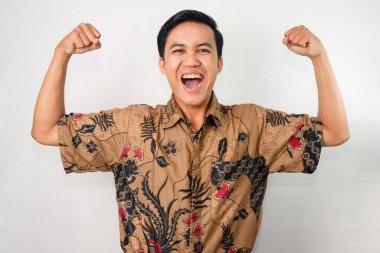 Açık ağızlı, batik giyinmiş ve iki kolunun kaslarını gösteren jestler yapan Asyalı bir adam.