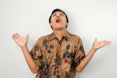 Şaşırmış bir ifadeyle etkileyici bir Asyalı adam, batik giyiyor ve elleri yukarı dönük bir jest yapıyor.