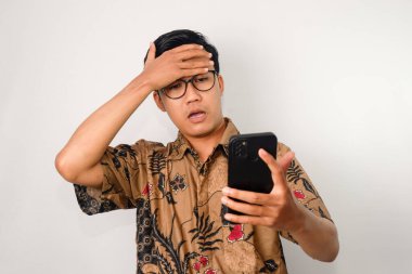 Şok olmuş suratlı, batik tişörtlü ve gözlüklü, alnına tokat atan Asyalı bir adam.