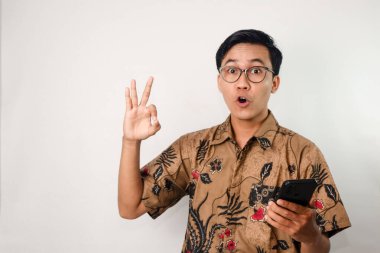 Neşeli bir suratı olan Asyalı bir adam batik gömlek ve gözlük takıyor, elinde akıllı telefon, diğer elinde de güzel bir jest yapıyor.