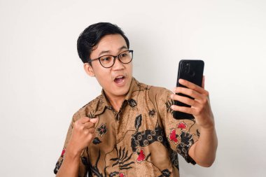 Mutlu bir suratı olan Asyalı bir adam batik gömlek ve gözlük takıyor, akıllı telefonuna bakıyor ve başarısını kutluyor.