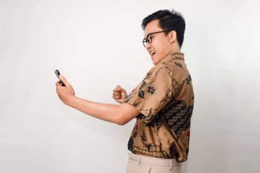 Gözlüklü, mutlu bir suratı olan, batik tişörtlü akıllı telefonuna bakıp başarısını kutlayan Asyalı bir adam.