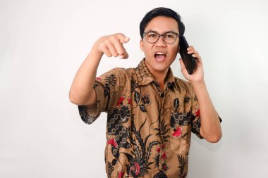 Kızgın yüzlü, gözlüklü ve batik tişörtlü Asyalı bir adam birini arıyor ve eli kamerayı işaret ediyor.