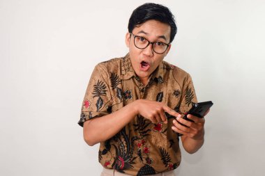 Ciddi ve komik yüzlü, gözlüklü ve batik gömlekli bir Asyalı akıllı telefonunu işaret ediyor.