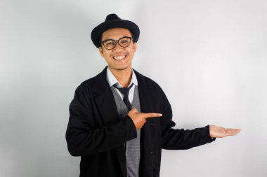 Asyalı bir adam mafya gibi giyinmiş, gözlük takıyor ve şapka takıyor. Dost canlısı bir yüzü var. Elleri ürünü işaret ediyor.