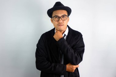 Mafya gibi giyinmiş, gözlüklü ve şapkalı Asyalı bir adam çenesinde garip bir ifadeyle el hareketi yapıyor.