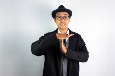 Mafya gibi giyinmiş, gözlüklü ve şapkalı Asyalı bir adam paniklemiş bir ifadeyle el hareketi yapıyor.