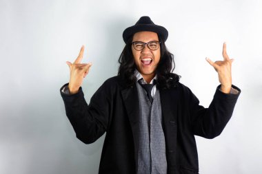 Mafya gibi giyinmiş uzun saçlı Asyalı adam gözlük takıyor ve şapka takıyor. Heyecanlı ifadeyle metal ya da kaya hareketleri yapıyor.