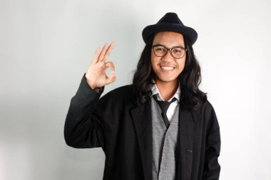 Uzun saçlı Asyalı adam mafya gibi giyinmiş gözlüklü ve şapkalı, gülen yüzlü iyi bir jest yapıyor.