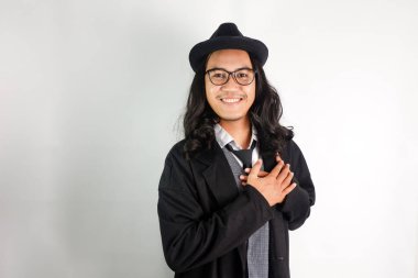 Uzun saçlı, mafya gibi giyinmiş, gözlüklü ve şapkalı Asyalı bir adam, gülümseyen yüzüyle göğsünü tutarak bir jest yapıyor.